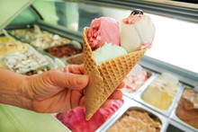 Female Hand Is Holding A Large Strawberry, Mint, Vanilla With Sour Cherry Ice Cream In Waffle Cone. Ice Cream Fridge With Steel Service Containers In Background. Worker In Ice Cream Shop. Pastry Shop.