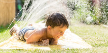Boy Cooling Down With Garden Hose, Family In The Background On A Hot Summer Day