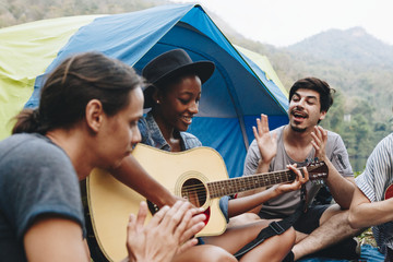 group of young adult friends in camp site playing guitar and ukelele and singing together outdoors r
