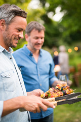 Wall Mural - in a summer evening,  two men  in their forties prepares a barbecue for  friends gathered around a table in the garden