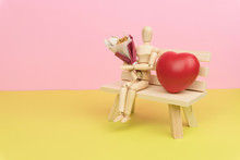 Love And Relationship Concept, Wood Figure Holding A Bouquet Of Dried Flower And Sitting On The Wooden Bench With A Red Love Heart Symbol