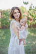 © Serenkonata - A beautiful young mother with a baby in her arms is standing in the garden.