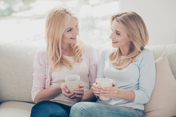 Wall Mural - Trendy charming pretty cheerful mother and daughter family with one parent adult child sitting in livingroom indoor holding mugs with tea cacao beverage looking at each other enjoying free time