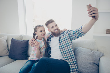 Poster - Cute girl with bearded stylish father in casual outfit checkered shirts sitting on couch in house embracing shooting self portrait on front camera showing peace symbol holding cell smart phone in hand