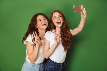 Portrait of two joyful women with ginger hair taking selfie on smartphone and showing victory sign, isolated over green background