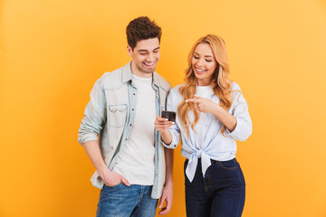 Photo of positive people man and woman smiling while gesturing at mobile phone, isolated over yellow background