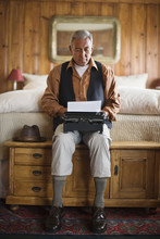Senior Adult Man Sitting At The End Of A Bed Using A Typewriter.