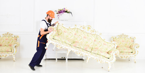 Wall Mural - Loader moves sofa, couch. Courier delivers furniture in case of move out, relocation. Delivery service concept. Man with beard, worker in overalls and helmet lifts up sofa, white background.