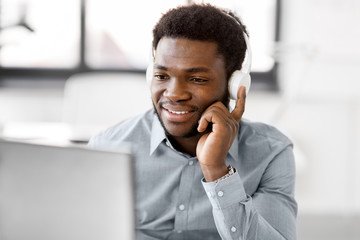 business, technology and people concept - happy african american businessman with headphones and computer listening to music at office