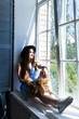 © Inna Vlasova - Long hair woman with a hatsits on the windowsill at the window by the warm glow of the sun shining indoors. Relaxing, thinking, enjoying in the living room. Summer girl portrait.