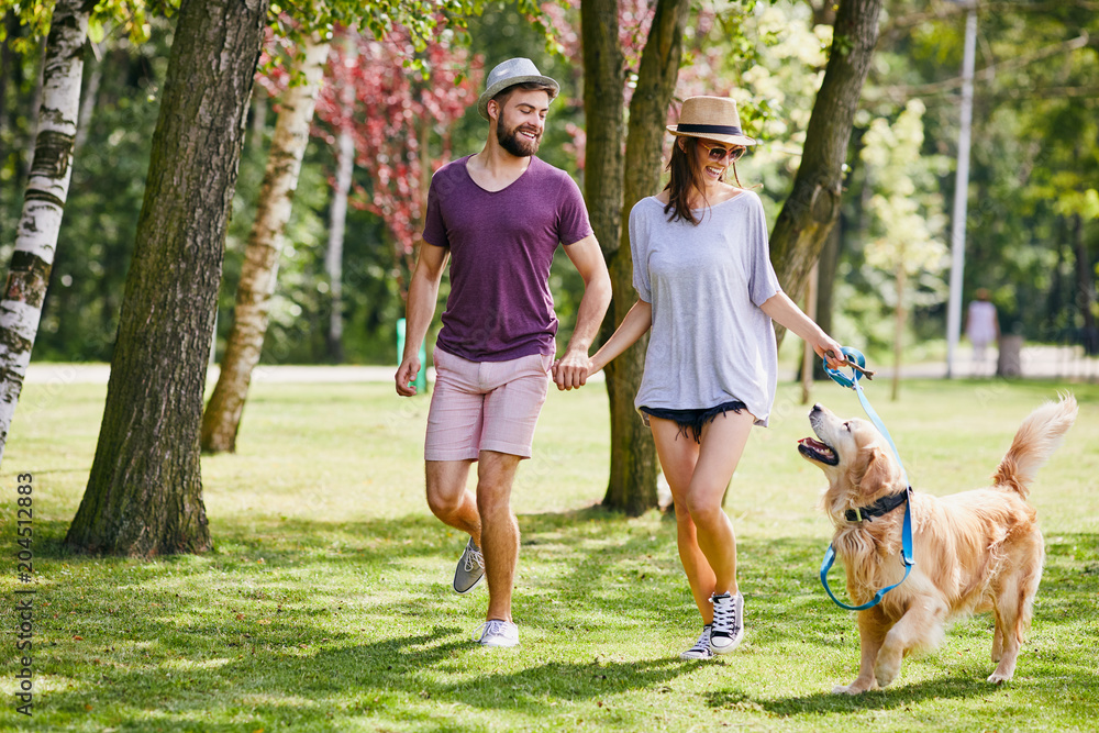 Young couple walking their dog together in the park during summer - obrazy, fototapety, plakaty 