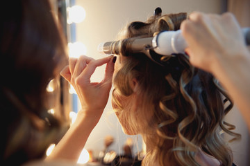 Hairstylist make beautiful curls with curler on light-brown with blonde client's hair. Creating a luxurious evening hairstyle with ringlets of medium length.