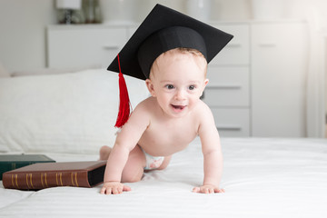 funny image of adorable baby boy in diapers wearing graduation cap crawling on bed