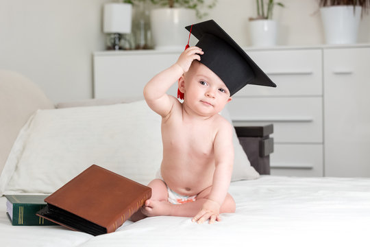 Cute baby genius baby in university graduation cap rubbing forehead and looking in camera. Concept of early child education