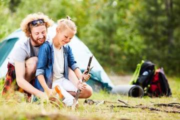 Wall Mural - Young man and his son sharpening sticks with axe while going to prepare food on campfire