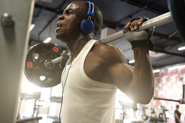 Wall Mural - A black muscular guy in a white T-shirt in blue headphones holds a barbell on his shoulders.