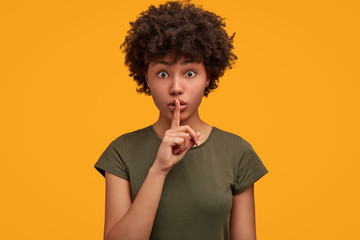 Wall Mural - Isolated shot of surprised young African American female gestures indoor, keeps fore finger on lips, wears casual t shirt, poses against yellow background. Please, don`t speak loudly! Hush sign