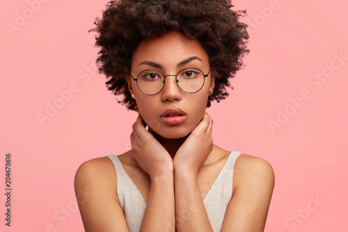 Cuadros en Lienzo Close up portrait of attractive dark skinned female has bushy Afro hairstyle, looks directly into camera, keeps hands on neck, isolated over pink background