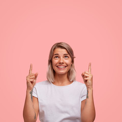 Wall Mural - Indoor studio shot of happy female hipster with pleasant gentle smile, has tattooed arms, indicates at blank copy space for your advertisement, isolated on pink background. Optimistic woman advertises