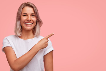 Wall Mural - Horizontal portrait of beautiful cheerful Caucasian female with braces, has broad smile, indicates at blank copy space for your promotional content, smiles joyfully. People, advertisement concept