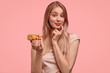 Charming young lovely female holds sweet doughnut, proposes you to eat delicious dessert, can`t refuse tasty eating, isolated over pink background. Gorgeous cute woman with donut. Junk food.