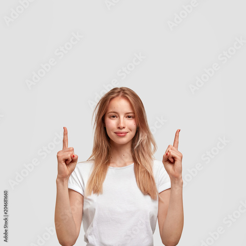 Fotografia Vertical shot of Caucasian beautiful female has attractive look, indicates upwards with fore fingers, wears white clothes in one tone with background to show monochrome