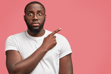 Wall Mural - Waist up portrait of handsome attractive plump African American male looks in stupor at camera, wears spectacles and white casual t shirt, indicates at blank copy space for your advertisement