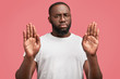 Middle aged black male shows stop gesture, keeps palms in front, has serious expression, wears casual t shirt, isolated on pink background. That`s forbidden! African American man rejects something