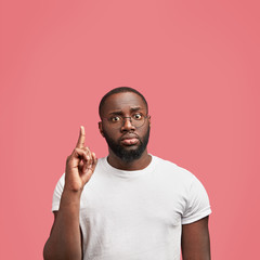 Wall Mural - Young serious plump African American male with brilliant idea in mind, keeps fore finger raised, wears round spectacles, poses against pink background with copy space up. Dark skinned man gestures