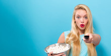 Young Woman Eating Popcorn And Holding A TV Remote Control On A Solid Background