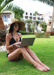 Sexy girl in a black bikini sits on a green lawn with a laptop on the hotel's territory. Find a tour. Video blog. Delicate shiny skin.
