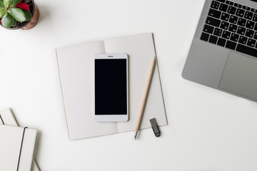 Top view of office workplace. White desk with copy space. Flat lay view on table with laptop, phone, flower, notebooks, flash drive and pencil. Creative designer concept.