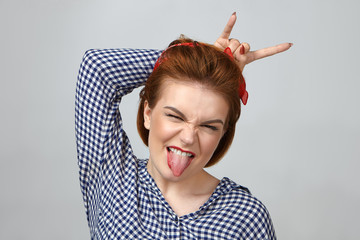 Wall Mural - People, lifestyle, emotions, fun and relaxation concept. Picture of stylish emotional young female in checkered shirt sticking out tongue and showing devil horns sign while enjoying live rock concert