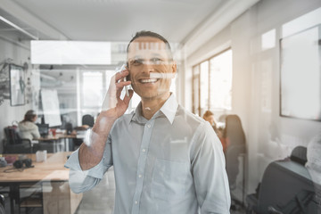 Wall Mural - Portrait of happy male talking by phone while looking at window. Cheerful worker with gadget concept