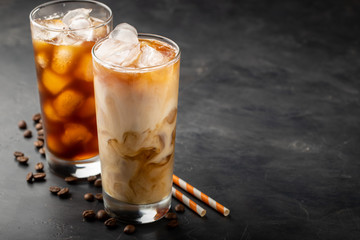 Ice coffee in a tall glass with cream poured over, brown sugar and coffee beans. Cold summer drink on a dark background. With copy space