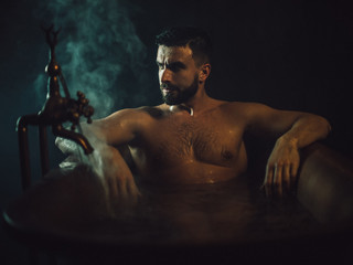 a handsome, muscular man with a beard lies in a bronze bathroom. steam coming from the hot water. he