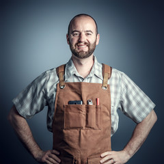  studio portrait of a caucasian carpenter wearing a classic leather apron. arms resting on the sides and serene and confident expression.