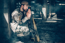 Spiritual Man Is Sitting In Squat Position And Praying. He Is Keeping His Eyes Closed And Holding Hands Together Close To Face. Also There Is A Rifle Near His Knees.