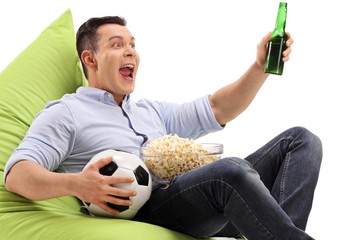 Wall Mural - Young man with a bottle of beer and a bowl of popcorn sitting on a beanbag and watching soccer on television