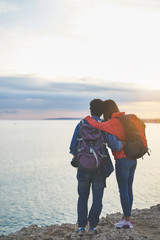 Wall Mural - Peaceful young man and woman are watching the sunset at the seaside. They are standing with backpacks and hugging