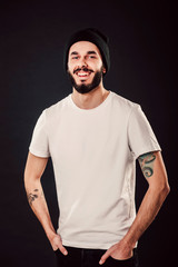 Wall Mural - Waist up portrait of serious stylish attractive man with thick beard looks confidently, dressed in casual white t shirt, isolated over black background. Young hipster in cap with tattoo. Mock up
