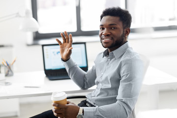 Poster - business and people concept - african american businessman with coffee sitting at office table and waving hand