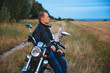 © lkoimages - Young biker wears black Leather Jacket and jeans, sits on his black old motorbike, holds white helmet and looks on beautiful soft sunset