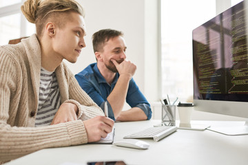 Side view portrait of two contemporary program developers writing code sitting at desk using computer and collaborating on startup project in modern office, copy space