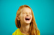 Loud and strong laughter of little girl with red hair, wearing yellow t-shirt, a shot of child on isolated blue.