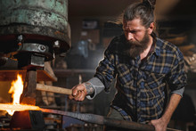 Real Brutal Blacksmith Works In A Workshop Mechanical Hammer With A Red-hot Iron. Portrait Of A Profession.