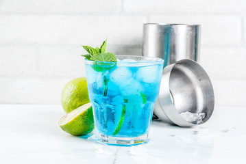 Colorful summer beverage, iced blue lagoon alcohol cocktail drink with lime and mint, white marble background copy space