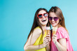 © proimagecontent - Two pretty happy smiling girls dressed in colorful t-shirts and sunglasses have fun with icecream on blue background, carefree youth concept