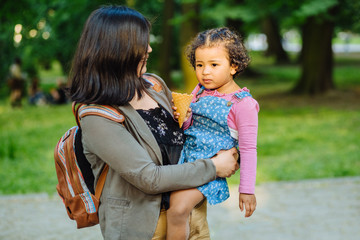 Mexican mixed race mother with dark skinned hungry hispanic toddler girl eating ice cream during a walk outdoors.