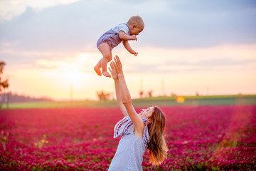 Wall Mural - Young mother, embracing with tenderness and care her toddler baby boy in crimson clover field, smiling happily